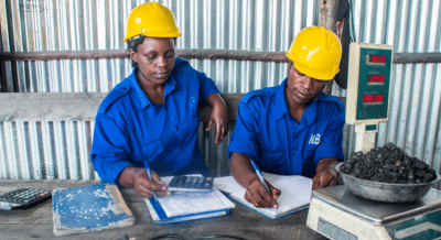 Two people in hard hats and overalls at a desk weighing and recording minerals