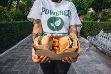 Person holding cardboard box with chips and burgers, wearing a t-shirt which reads ' Powered by Plants'