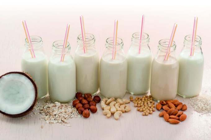 Small glass bottles of plant milks with the different raw ingredient in front e.g. nuts, oats